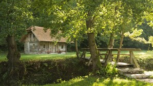 Une maison de campagne en pierre, dotée d’un toit en pente, entourée d’une végétation luxuriante et accessible par un pont en bois.