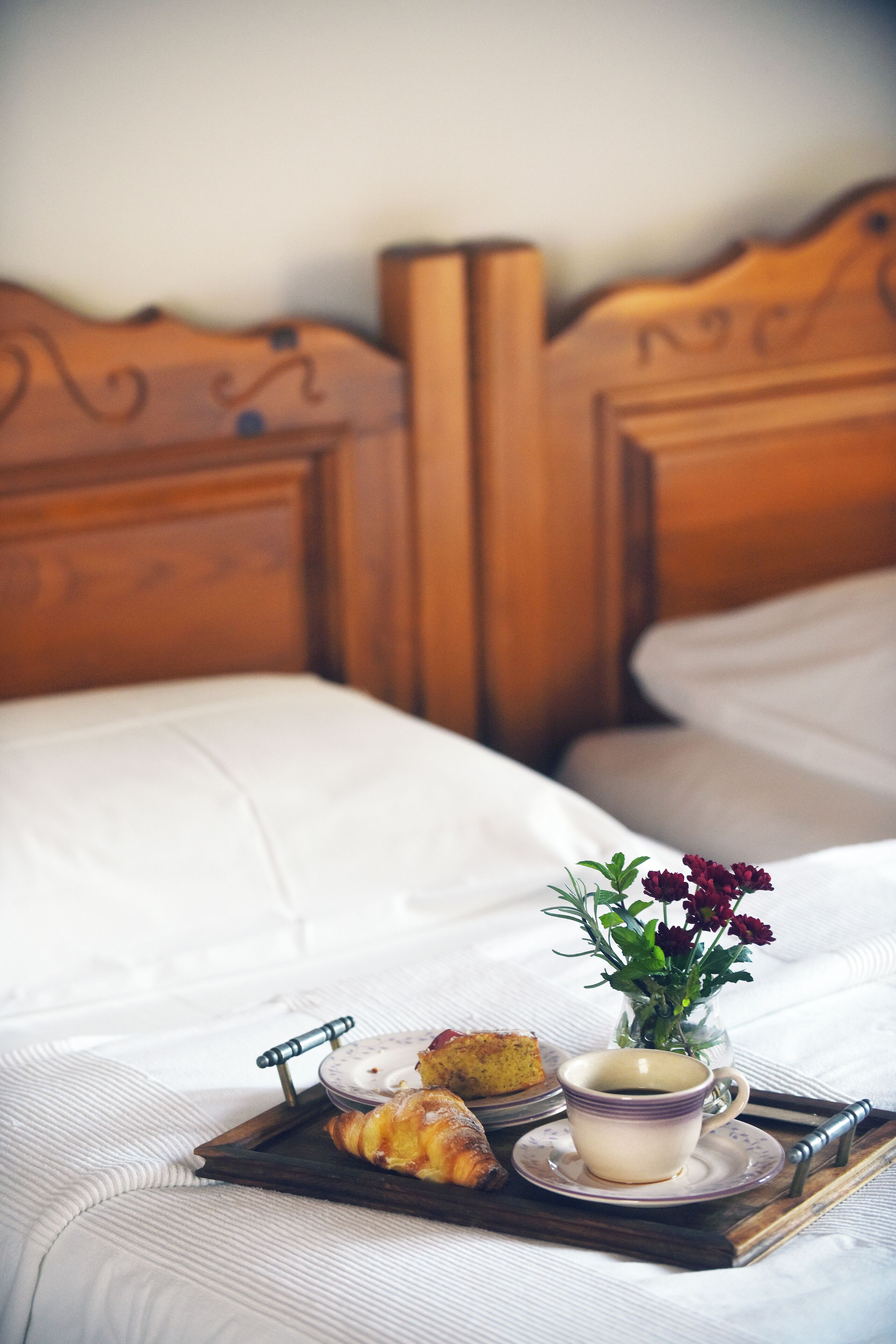 A neatly made bed with a tray of pastries, a cup of coffee, and a vase of flowers on a wooden headboard.