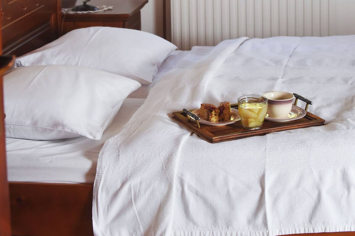 A bedroom with a wooden bed, white bedding, a bedside table with a lamp, a tray with breakfast items, and a window with curtains.