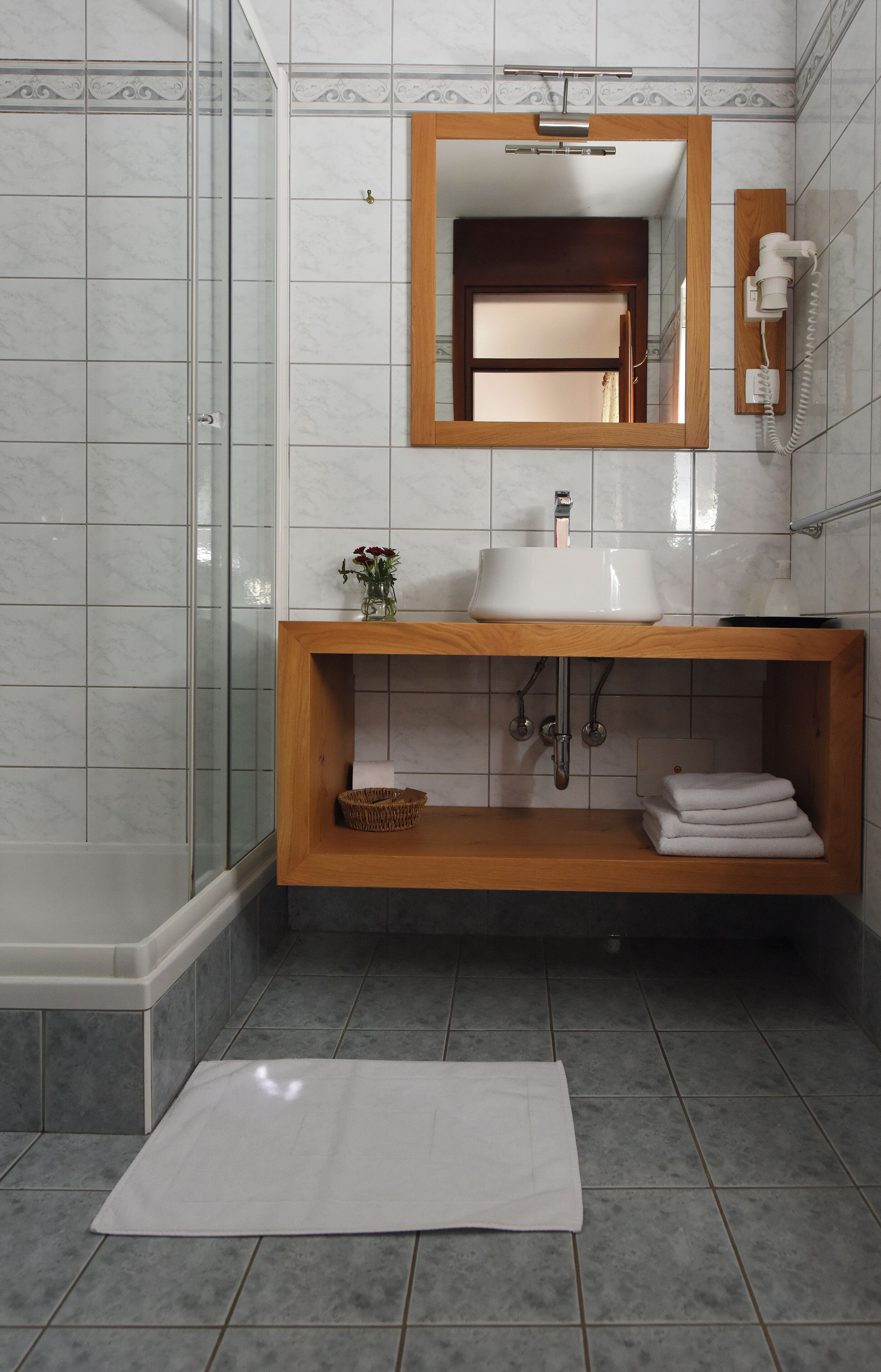 Un baño con cabina de ducha de vidrio, tocador de madera con lavamanos y espejo, y una alfombra de baño blanca en el suelo.