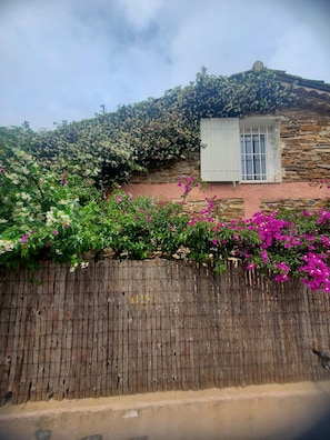 A rustic stone building with a wooden fence, a small window, and flowering plants.