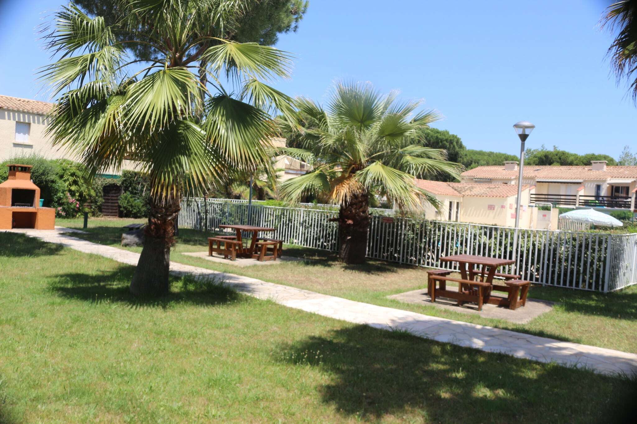 Un espace extérieur ensoleillé agrémenté de palmiers, de tables de pique-nique et d’une clôture blanche.