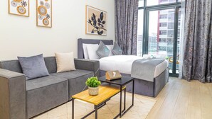 A modern bedroom with a grey sofa, a bed with a grey headboard, a wooden coffee table with a plant, and a city view through large windows. - Bloomfields STU View Azizi Riviera (Dubai)