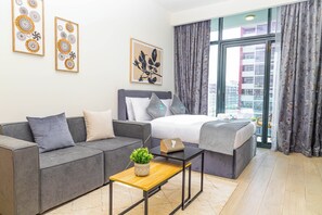 A modern bedroom with a grey sofa, a bed with a grey headboard, a wooden coffee table with a plant, and a city view through large windows.