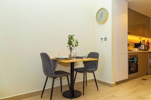 A dining area with a table set for two, a clock on the wall, and a kitchen in the background. - Bloomfields Studio - Azizi Riviera 36 - Meydan (Dubai)