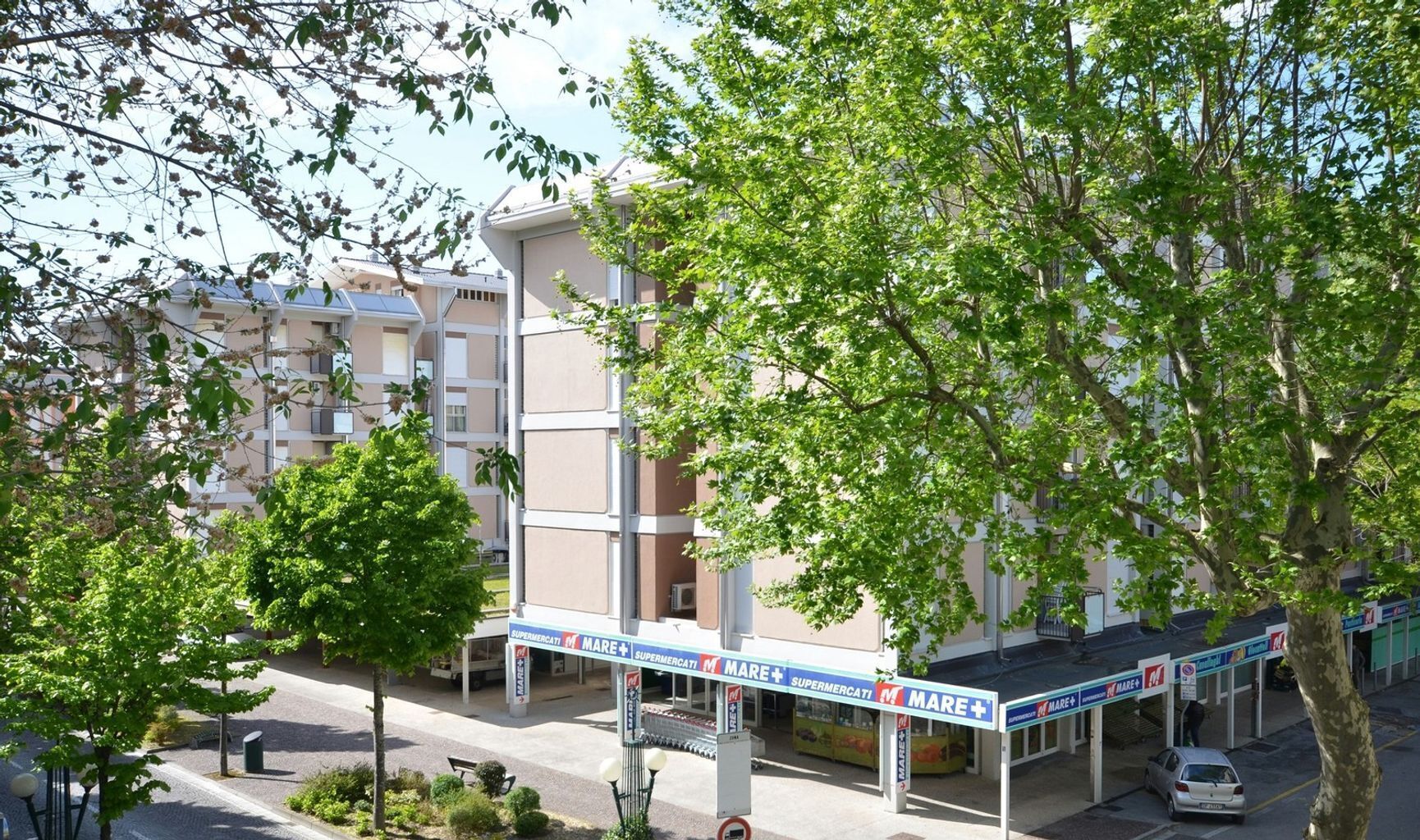A multi-story building with a covered outdoor area, possibly a parking lot or loading zone, with a sign indicating 'Mare+' and 'Supermercati'.