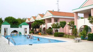Un espace piscine avec une terrasse carrelée, un petit bâtiment blanc doté d’un toit vert et une rangée de bâtiments roses avec des fenêtres en arc de cercle.