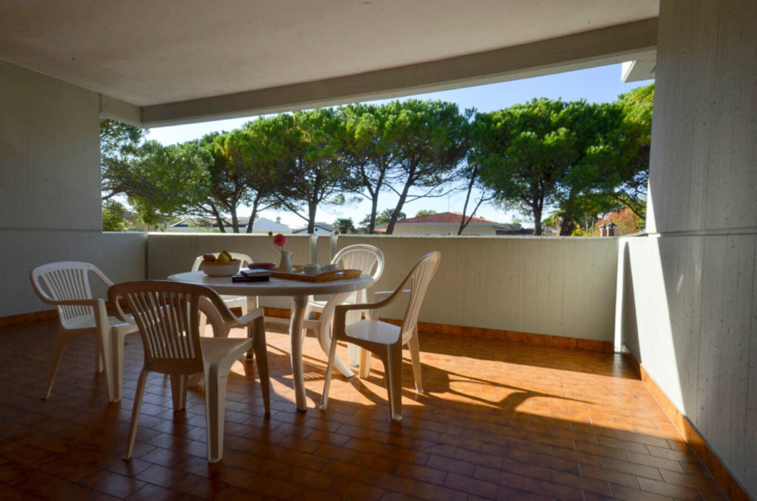 A covered patio with a round table and chairs, a bowl of fruit, and a view of trees and buildings.