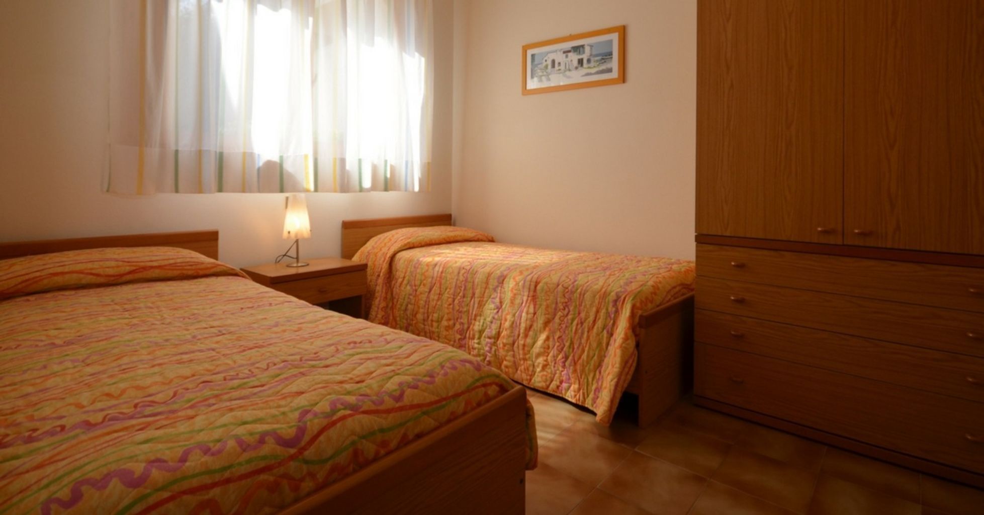 A bedroom with two beds, a nightstand, a lamp, a wooden wardrobe, and a framed picture on the wall.