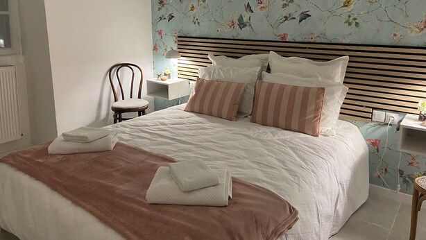 Un lit bien fait, avec des oreillers et une couverture, une table de chevet avec une lampe, et un papier peint à motifs floraux sur le mur.