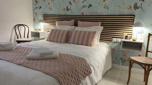 Un lit bien fait, recouvert d’une couverture rose, avec une tête de lit en bois et des oreillers à rayures. On y trouve une chaise et une table de chevet avec une lampe, placés à côté du lit.