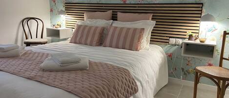 Een net opgemaakt bed met een roze sprei, een houten hoofdbord en gestreepte kussens. Er staat een stoel en een nachtkastje met een lamp naast het bed.