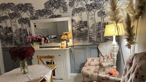 Un coin repas avec une table dressée pour deux personnes, un fauteuil à motifs floraux, un miroir et un mur orné d’une fresque représentant un arbre.