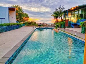 A rectangular swimming pool with clear blue water, surrounded by a paved deck and a low wall. There are palm trees and a building with large windows in the background. - Sunset Serenity Jomtien Resort (Sattahip)