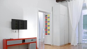 A room with a wooden floor, a red and grey desk with a television, a white door with a color-coded panel, and a window with white curtains. - Appartamento Casa toledo (Naples)