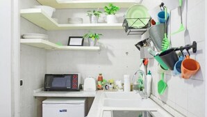 A compact kitchen with white cabinets, a built-in microwave, a sink with a glass basin, and various kitchen utensils and plants on shelves. - Appartamento Casa toledo (Naples)