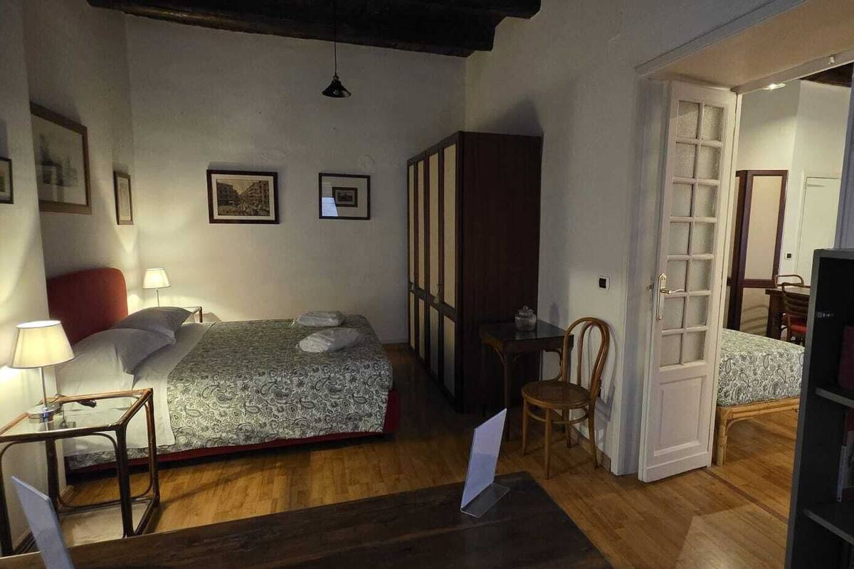 A small, cozy bedroom with a bed, bedside table, lamp, and a bookshelf.