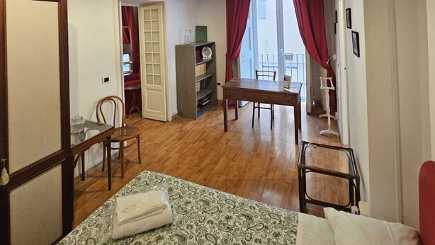 A room with a wooden ceiling, a bed with a patterned bedspread, a dining table with chairs, a bookshelf, and a window with red curtains. - Appartamento Casa toledo (Naples)