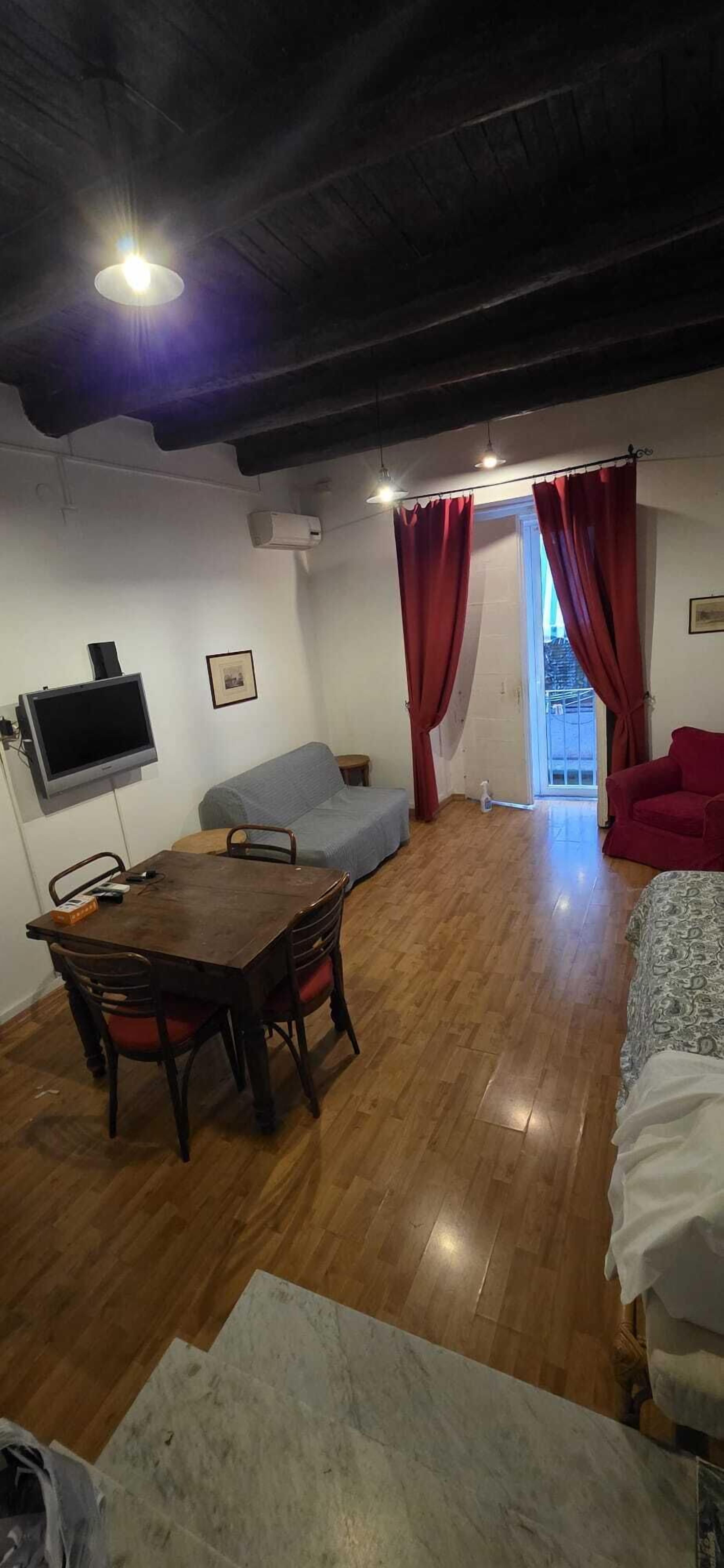 Une pièce avec un plancher en bois, une table à manger avec des chaises, un canapé, un lit, un téléviseur fixé au mur et une fenêtre avec des rideaux.