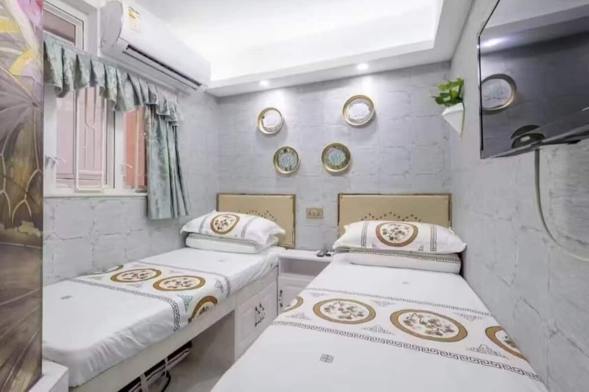 A small, compact room with two beds, a window with curtains, and a wall-mounted air conditioning unit.
