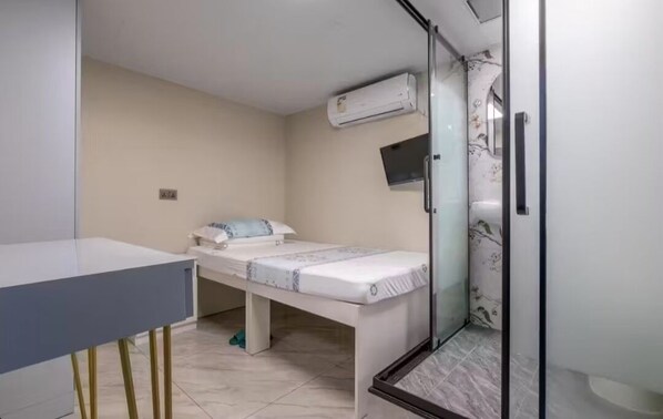 A small, modern bedroom with a single bed, a desk, and a shower area.