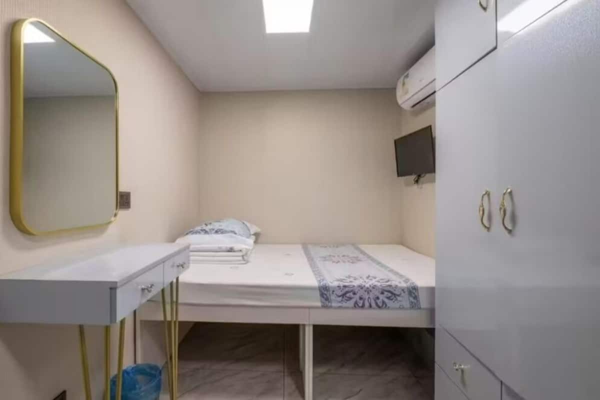 A small, compact room with a single bed, a bedside table, a mirror, a wardrobe, and a television on the wall.