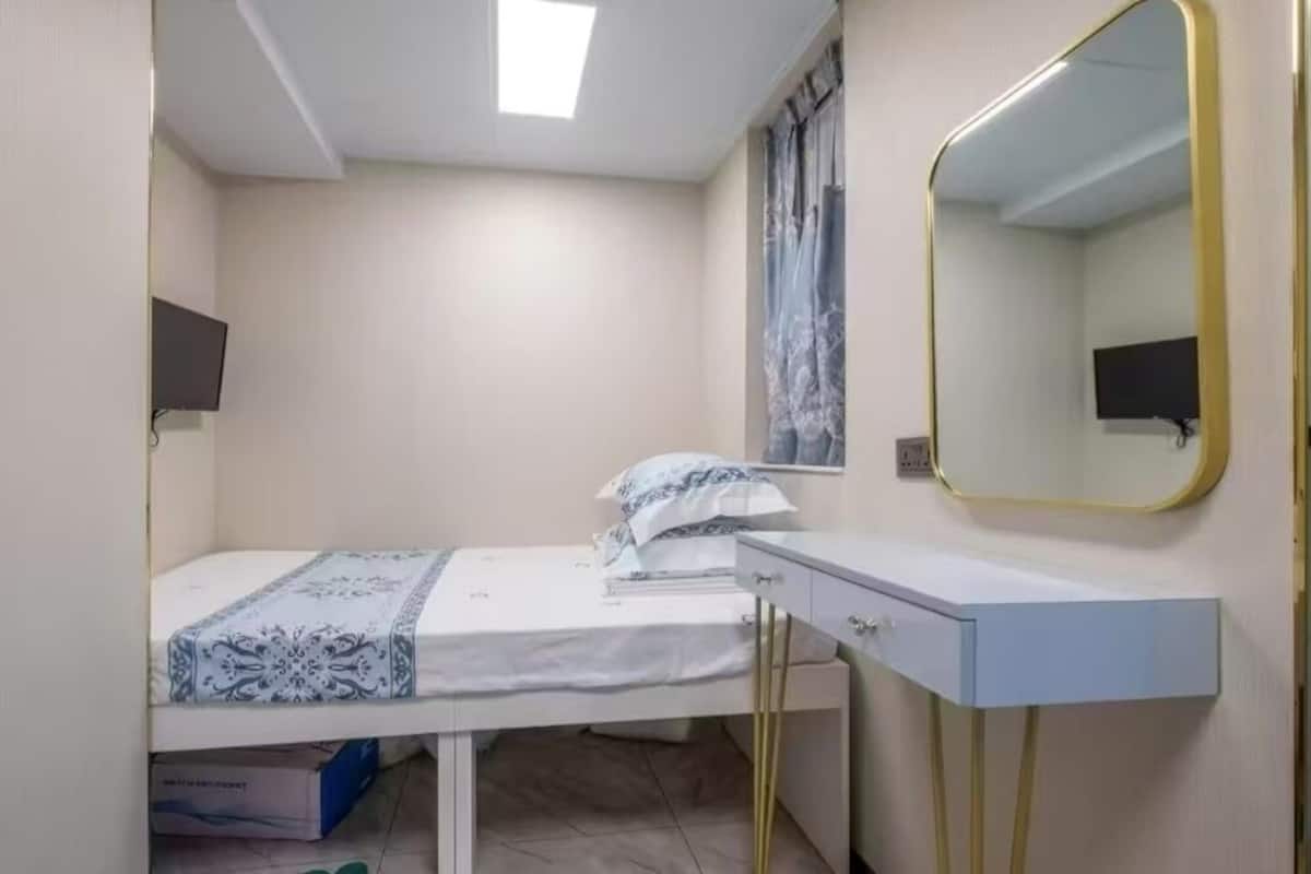 A small, neatly arranged bedroom with a single bed, a bedside table, a mirror, and a television mounted on the wall.