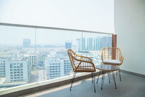 A balcony with two wicker chairs and a glass table, overlooking a cityscape with modern buildings.