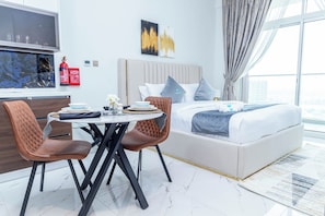 A modern hotel room with a bed, a dining table with chairs, a TV, and a fire extinguisher.