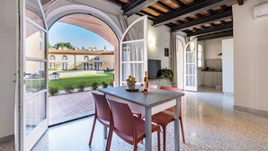 An open-plan living area with a dining table and chairs, a kitchen in the background, and a view of a courtyard with buildings and greenery.