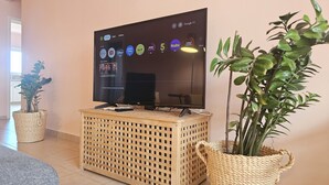 A flat-screen TV mounted on a wooden stand with a remote control, surrounded by two potted plants. - 07 LIM - Stay Velvet Apartment (Limassol)