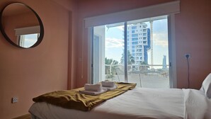 A hotel room with a bed, a window with a view of a building, a round mirror, and a power outlet. - 07 LIM - Stay Velvet Apartment (Limassol)