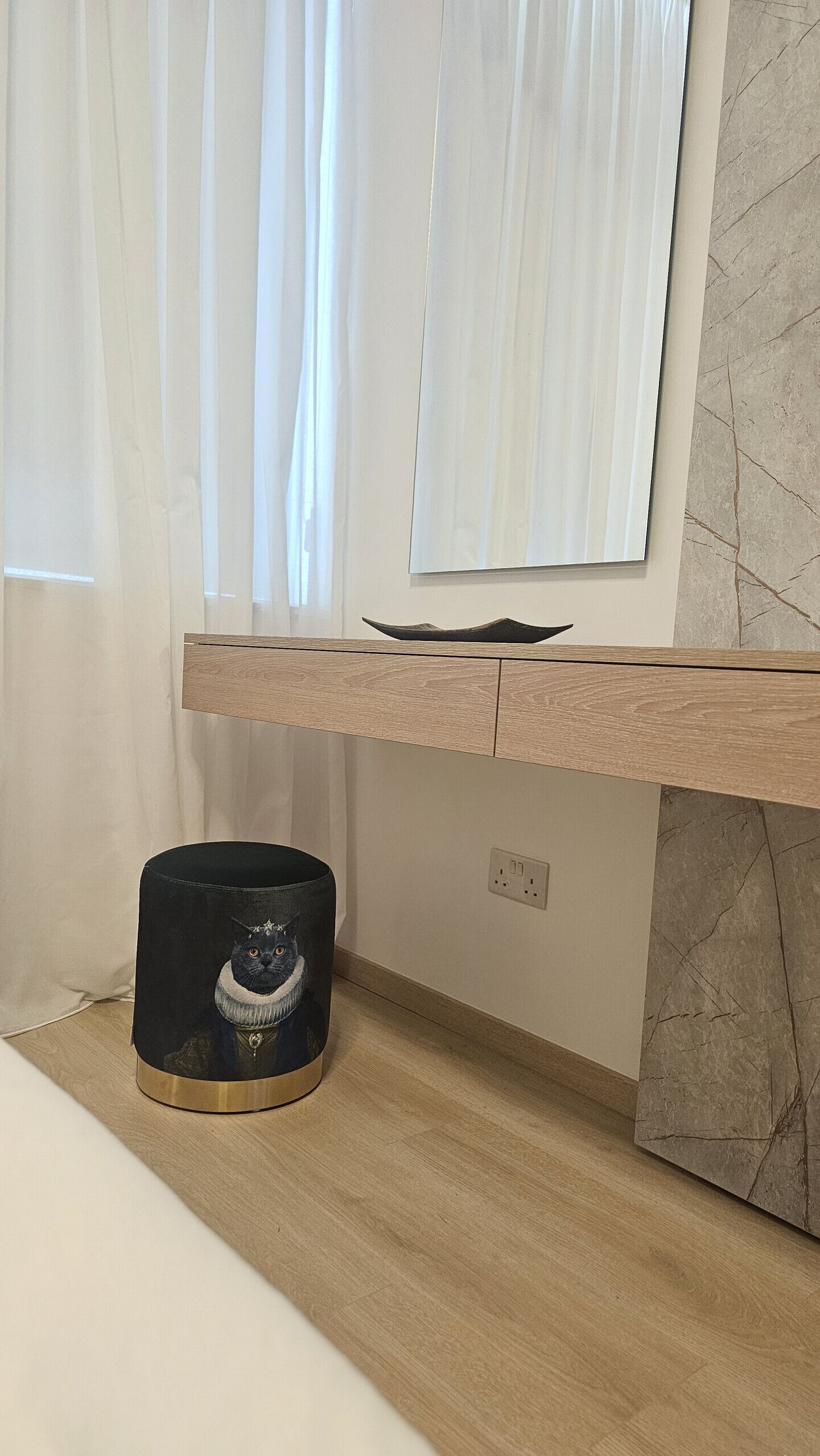 A room with a wooden desk, a black cylindrical object with a cat design, a marble wall, and a window with sheer curtains.