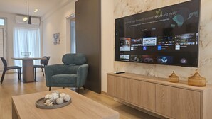 A modern living room with a large flat-screen TV displaying various streaming apps, a comfortable seating area with a sofa and chairs, a wooden coffee table with decorative items, and a marble wall.