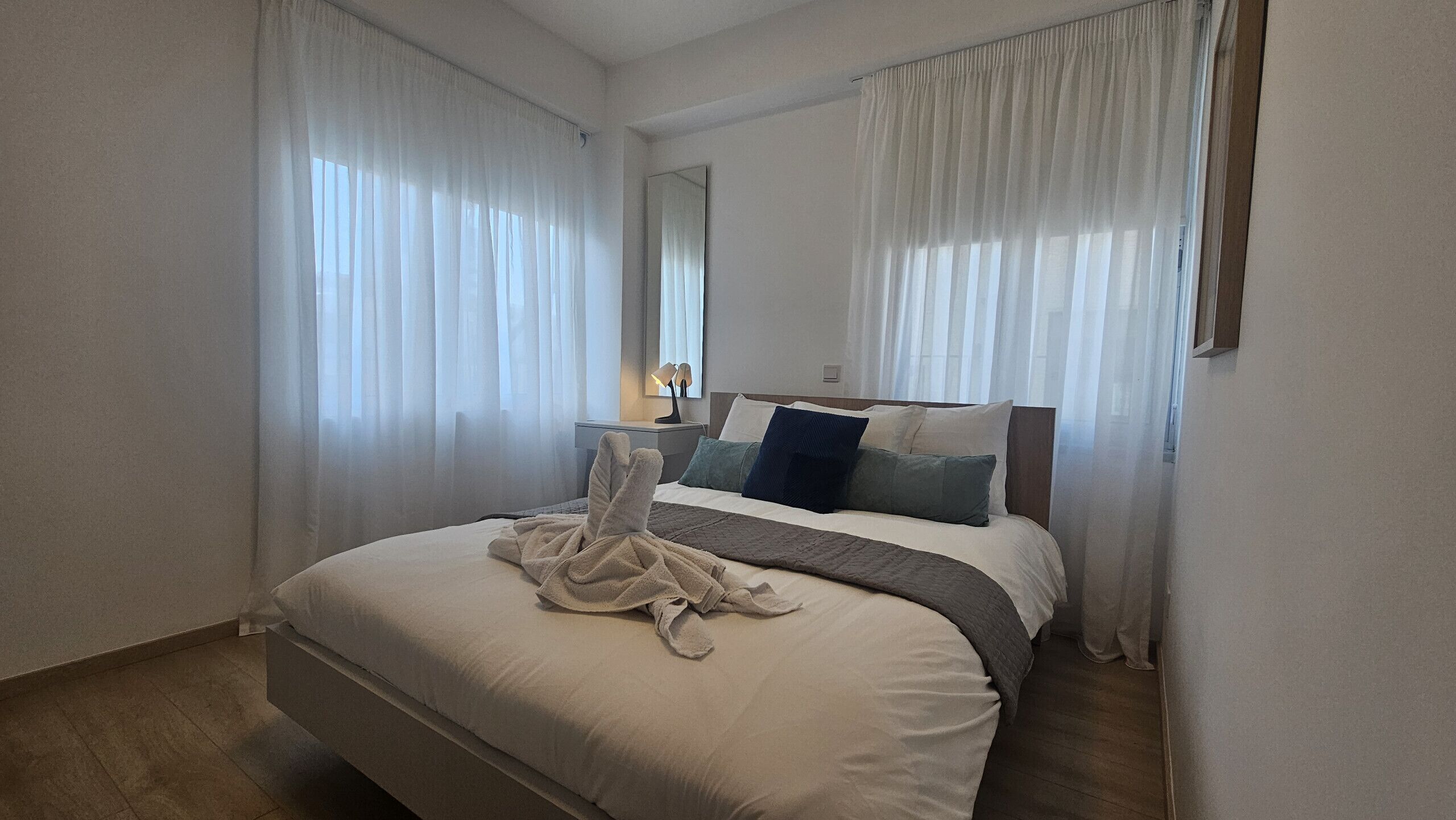 A hotel room with a bed, a nightstand, a mirror, and sheer curtains.