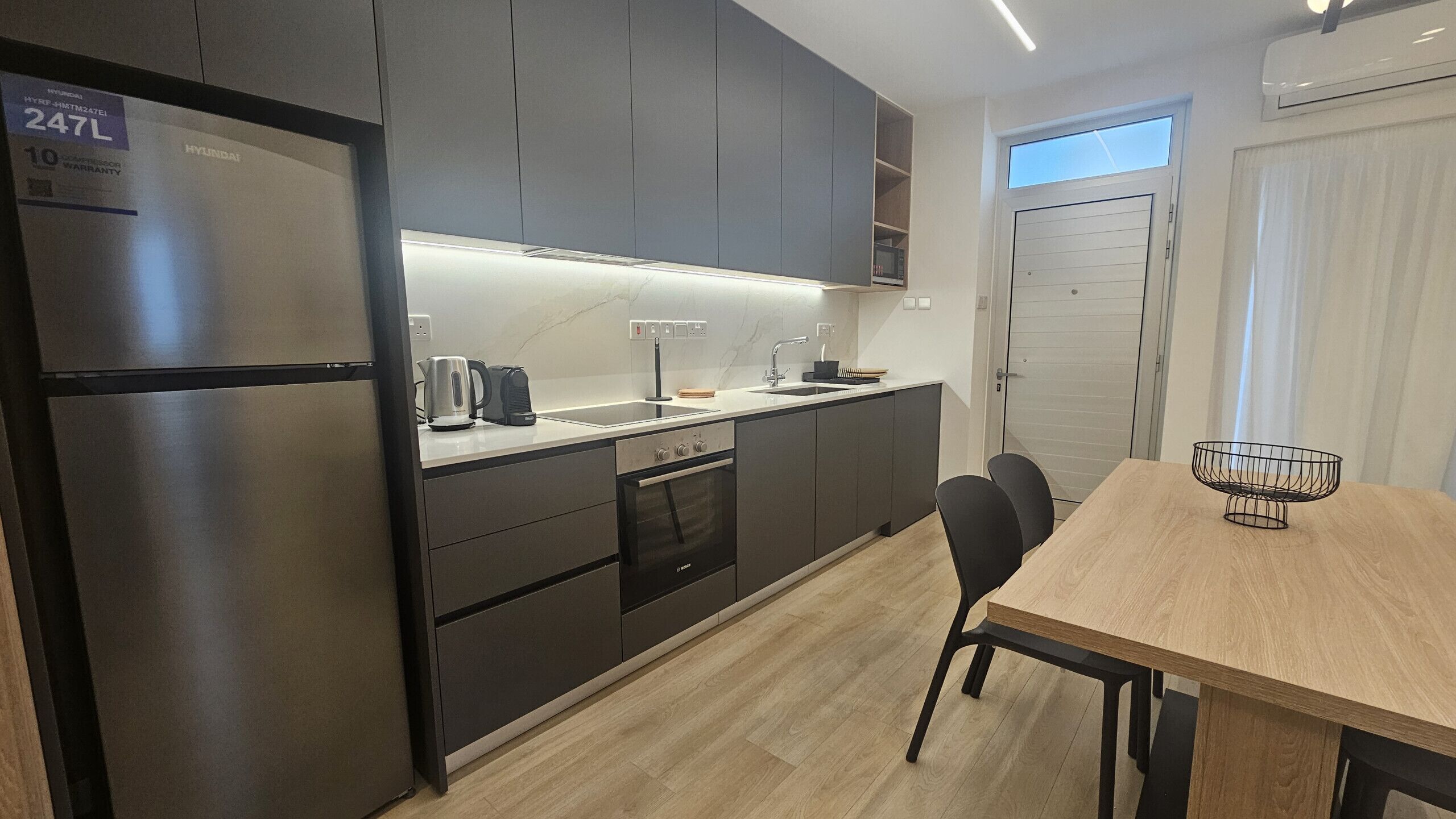 A modern kitchen with grey cabinets, a stainless steel refrigerator, and a wooden dining table with two chairs.