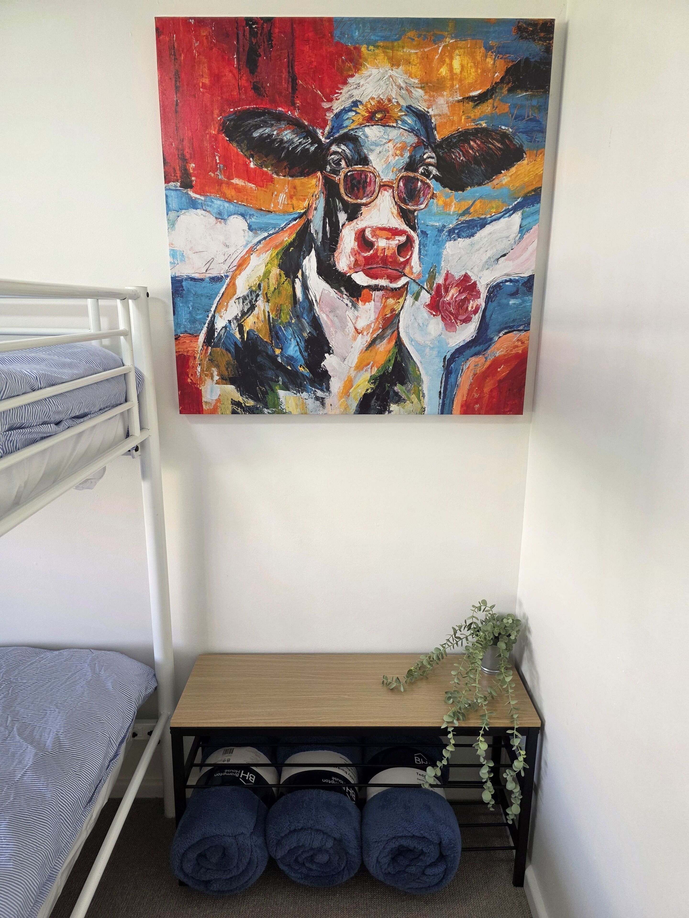 A bunk bed with a painting of a cow above it, a small table with towels, and a potted plant.