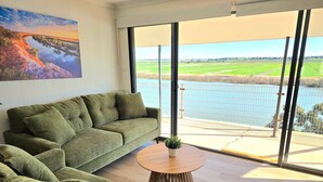 A modern living room with a green sofa, a round wooden table, and a large window offering a view of a body of water and fields. - RiverBed Rise (Tailem Bend)