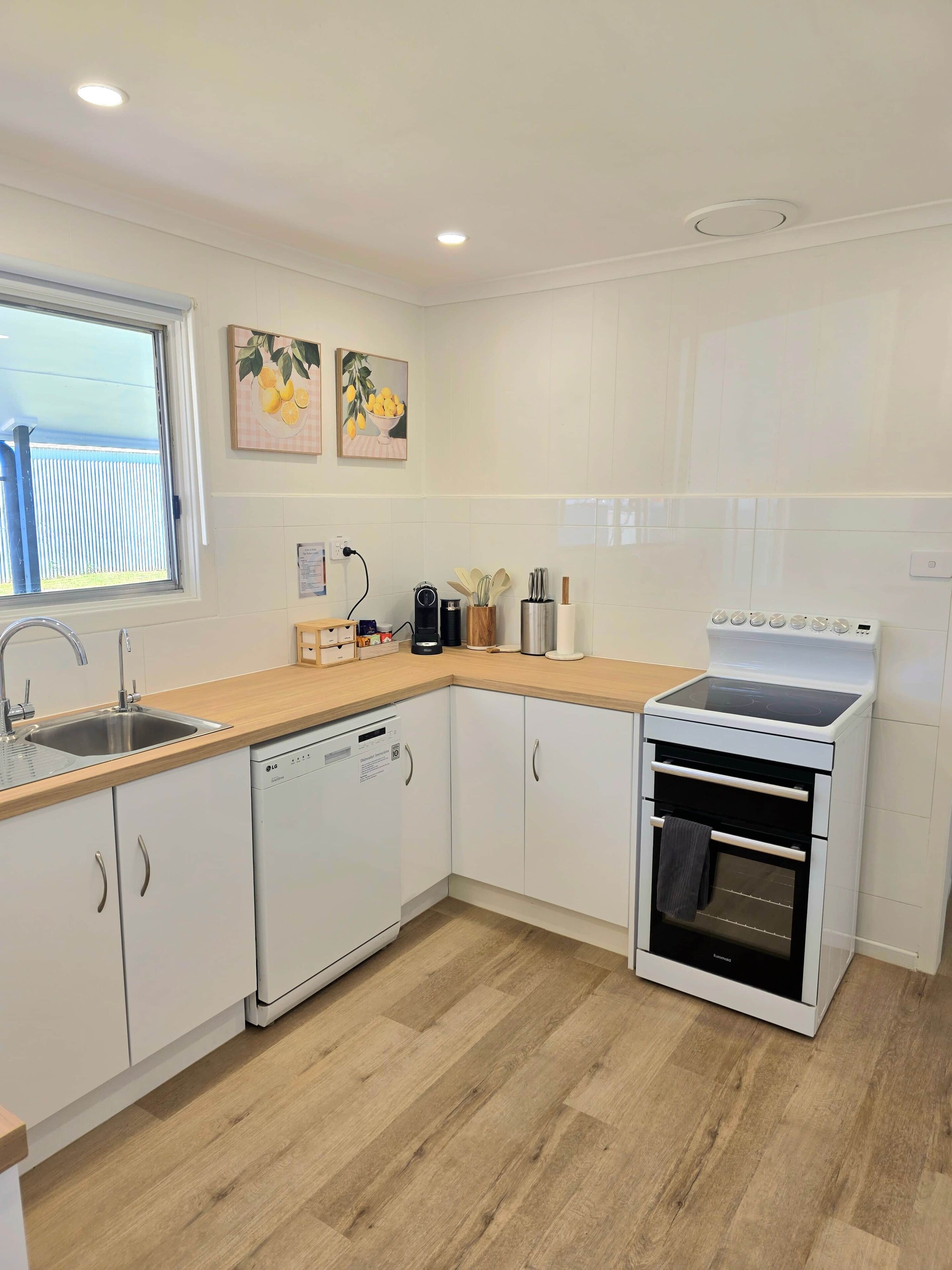 A modern kitchen with white cabinetry, a dishwasher, an oven, and a window with a view of a fence.