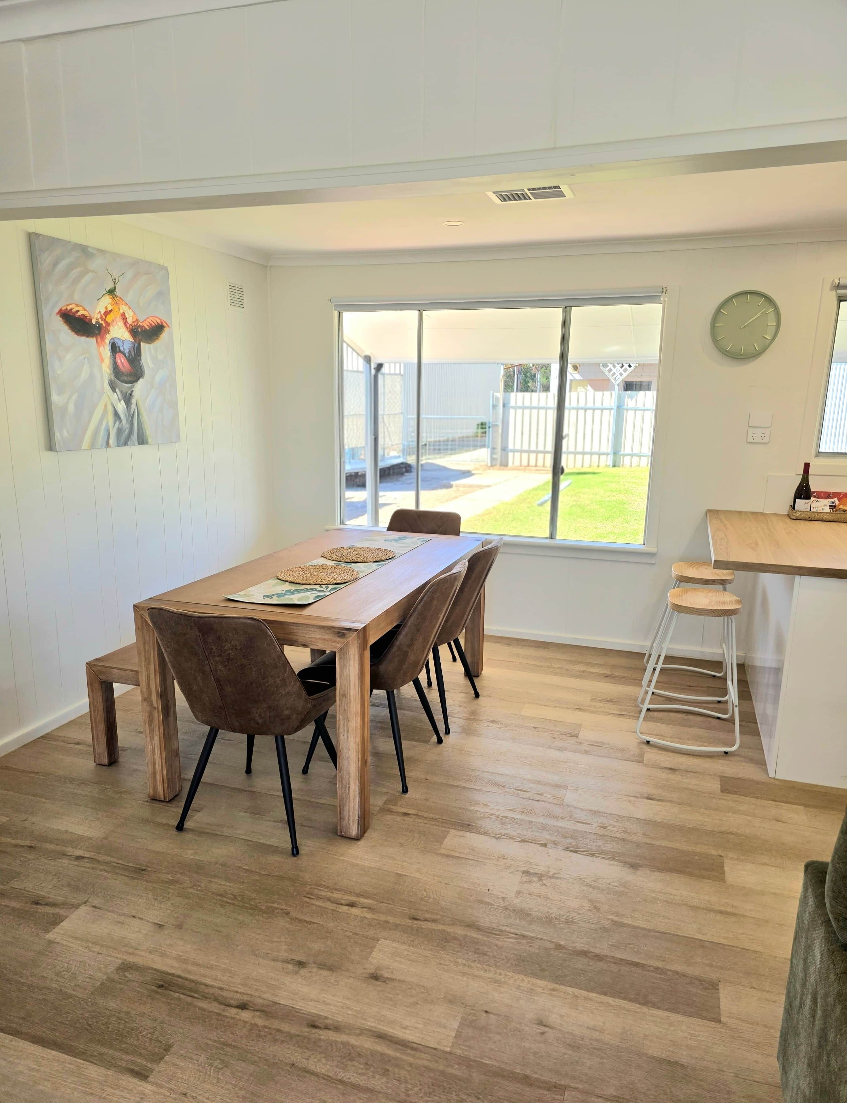 A dining area with a wooden table and chairs, a painting on the wall, and a view through a large window.