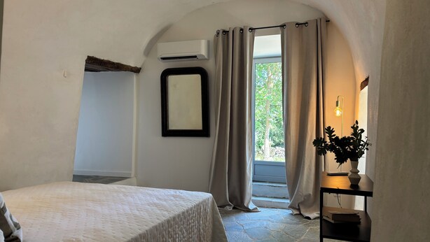 Un dormitorio con cama, aire acondicionado, espejo, ventana con cortinas, planta en maceta y escritorio con lámpara.