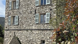 Un edificio de piedra con múltiples ventanas y contraventanas, rodeado de vegetación y un camino de piedra.