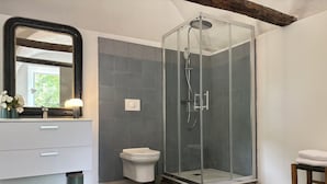 Un baño con ducha de vidrio, inodoro blanco, tocador con espejo y un pequeño taburete.