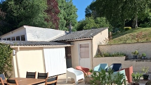 An outdoor patio with a wooden table and chairs, potted plants, and a small building with a door.