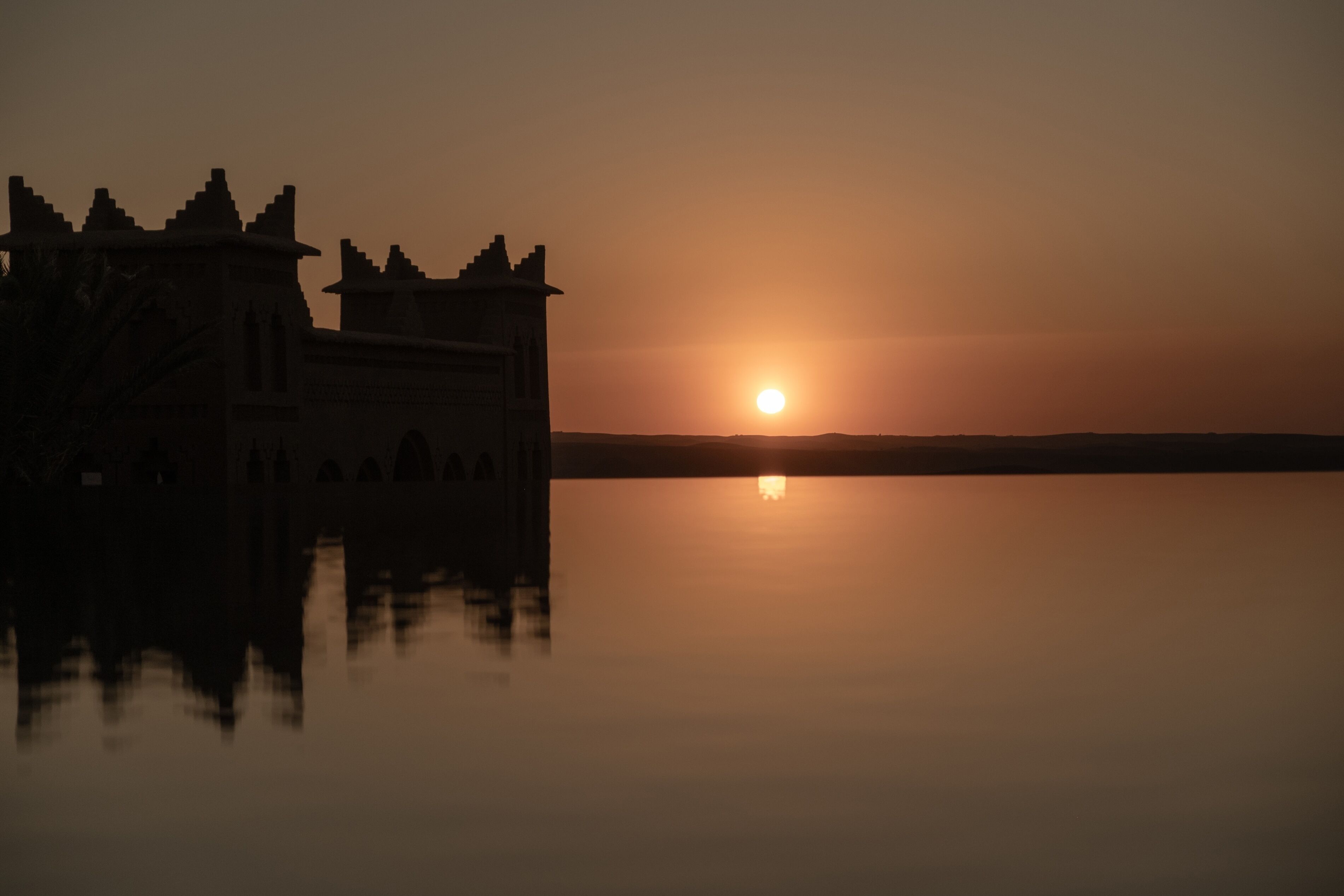 Une structure ressemblant à un château, dotée de tours et entourée de douves, qui se reflète dans les eaux calmes au moment du coucher du soleil.