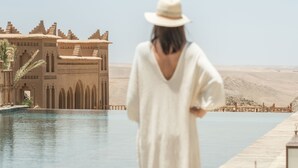Une personne vêtue d’une longue robe blanche se promène au bord d  ’une piscine, avec un paysage désertique et un grand bâtiment en arrière-plan.