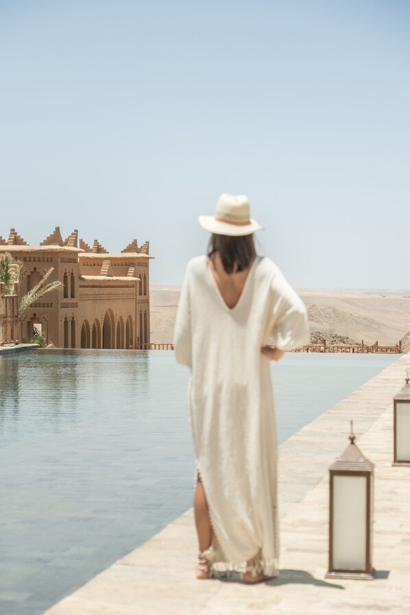 A person in a white robe walking along a poolside with a desert landscape and a large building in the background.