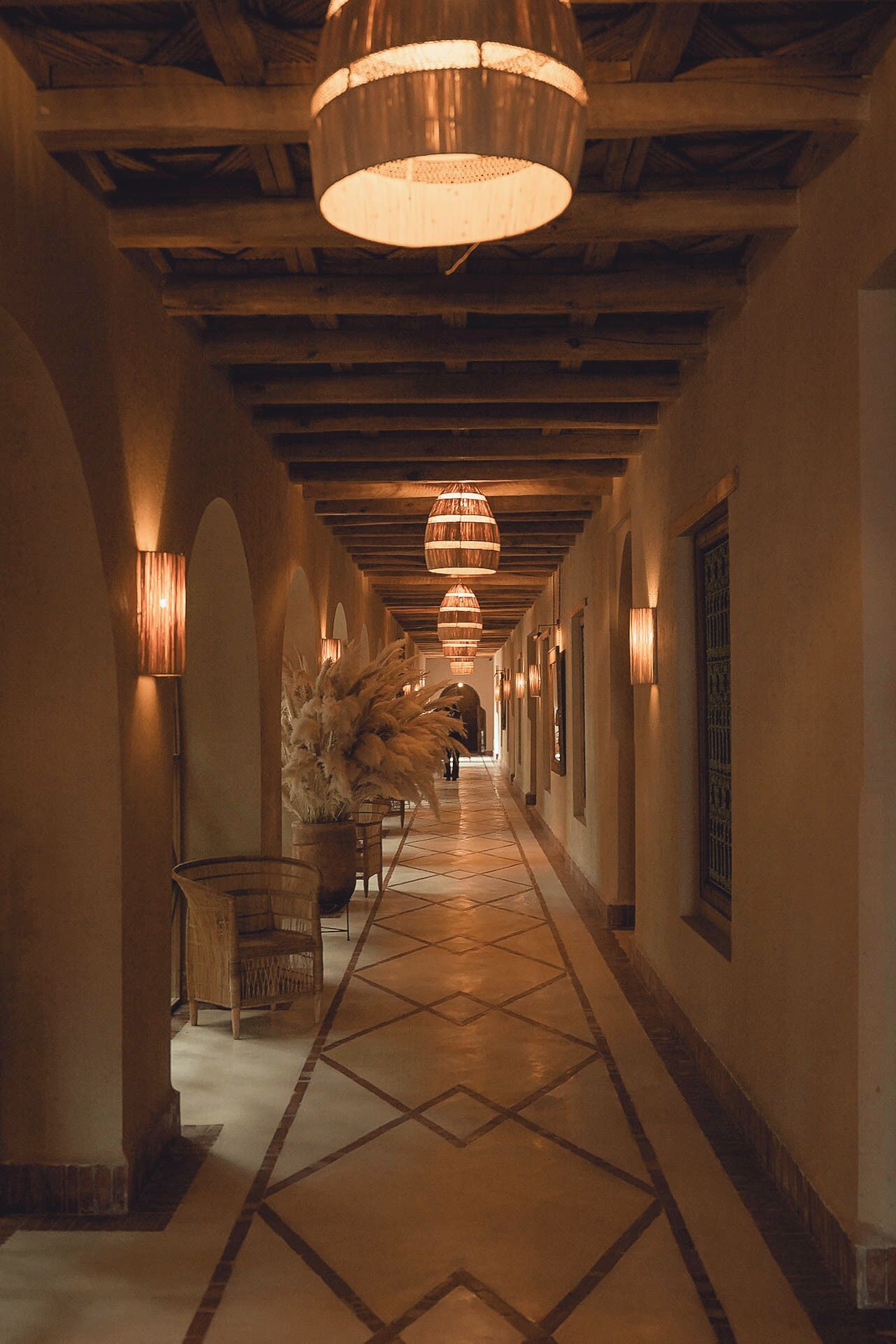 Un pasillo largo y estrecho con suelo de baldosas y vigas de madera en el techo. Varias lámparas colgantes y apliques de pared ofrecen una iluminación cálida.