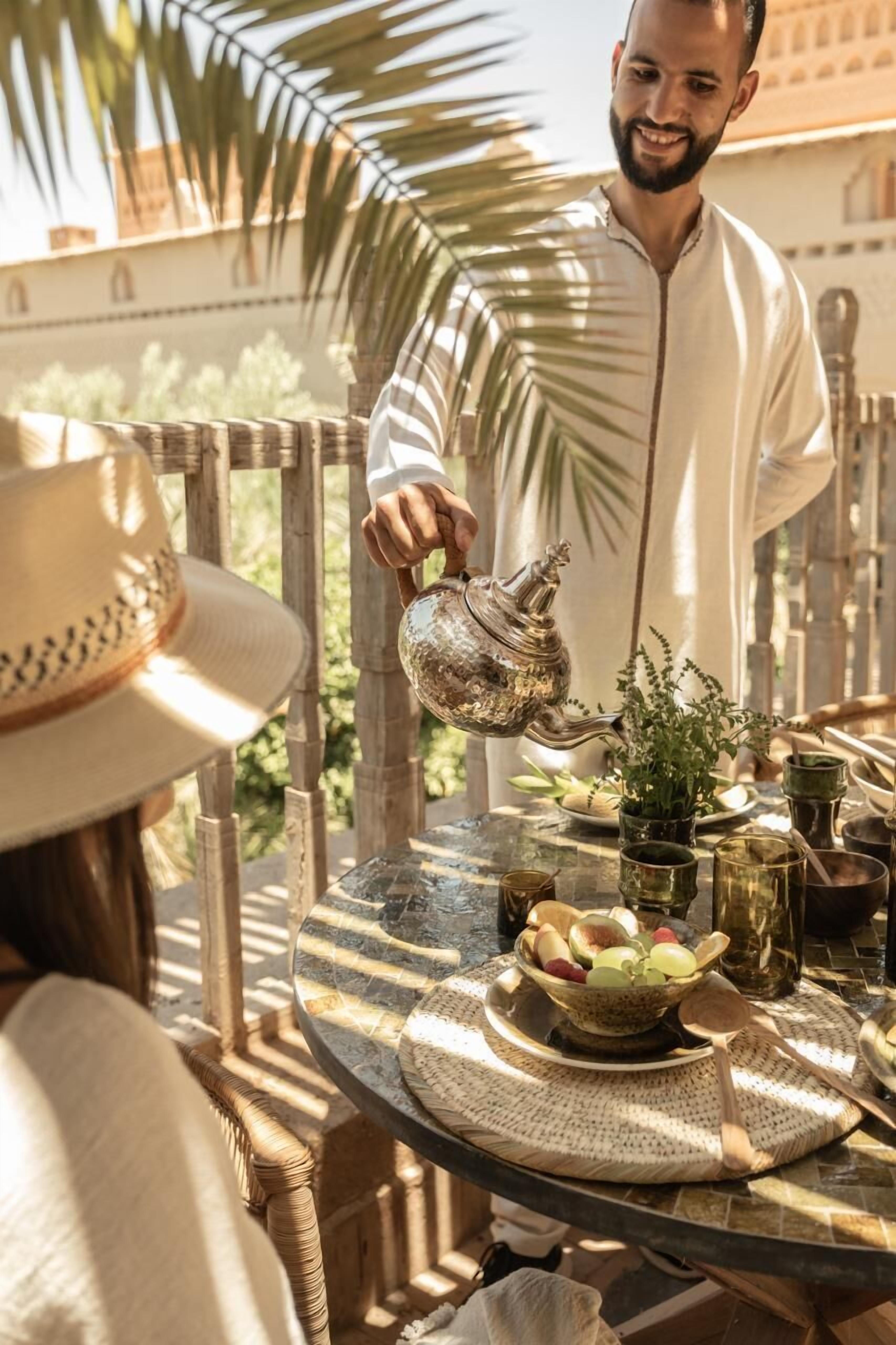 A person pouring water from a traditional metal teapot onto a table with a woven placemat, surrounded by various tea utensils and a bowl of fruit.