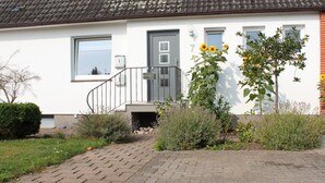 Ein Haus mit grauer Tür, ein kleiner Vorgarten mit einem Weg zur Tür und ein Busch mit gelben Blüten.